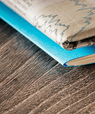 Close up of the corner of a folded newspaper and the corner of a blue paper folder, on top of a wooden table